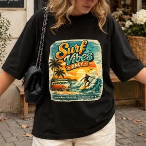 Peut inclure: T-shirt noir avec un motif coloré "Surf Vibes Only". Le design comprend un surfeur, un van vintage, des palmiers et un coucher de soleil. Le t-shirt porte également l'inscription "Catch the Wave - Ride the Tide - Live the Beach Life."