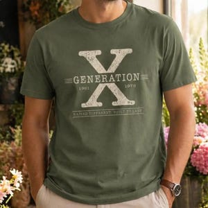 Op de afbeelding: Olijfgroen t-shirt met een witte afbeelding. De afbeelding toont een grote "X" met "GENERATION" erboven en "1961" en "1979" eronder. De tekst "RAISED DIFFERENT. BUILT TO LAST" staat onder de cijfers. Het shirt heeft korte mouwen.