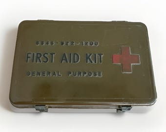 May include: An olive green metal first aid kit with the words "FIRST AID KIT GENERAL PURPOSE" in black lettering. A red cross is on the right side. The kit has a clasp closure and the number "6545-922-1200" is printed on the top.
