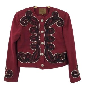 May include: A deep red jacket featuring a round neckline and long sleeves. The front and cuffs are decorated with dark brown braided cord trim and small white beads arranged in swirling patterns. Five silver-toned circular buttons fasten the front.