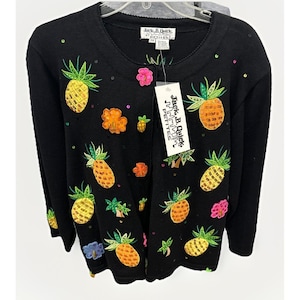 May include: Black cardigan sweater adorned with embroidered pineapples, flowers, and sequins. The sweater features a round neckline and long sleeves. The tag reads "Jack B. Quick Petites".