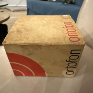 May include: A vintage, rectangular box with the word "ortofon" printed vertically in red and black. The box is beige with a red and white curved design on the corner. The box appears to be made of cardboard.