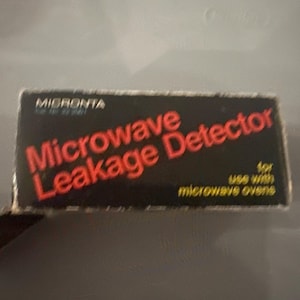 May include: A black box with the text "MIGRANTA" at the top. The words "Microwave Leakage Detector" are in red, with "for use with microwave ovens" below.