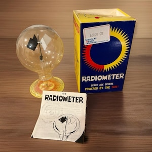 May include: A glass radiometer with a clear bulb containing black and white vanes on a yellow base, accompanied by a blue and yellow box labeled RADIOMETER SPACE AGE SPHERE POWERED BY THE SUN and a printed instruction manual with a diagram.