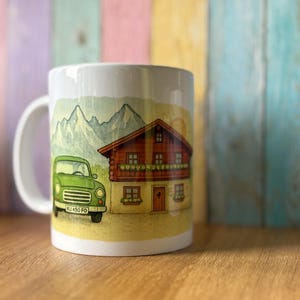 Könnte beinhalten: Weiße Keramiktasse mit einem Design eines grünen Oldtimers und eines Chalets mit rotem Dach. Der Hintergrund zeigt eine Bergkette. Die Tasse hat einen weißen Henkel und ist für Heißgetränke geeignet.