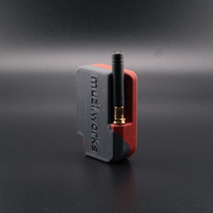Pode incluir: Um pequeno dispositivo preto e vermelho com uma antena preta. O dispositivo tem o texto "muzi.works" impresso no lado.