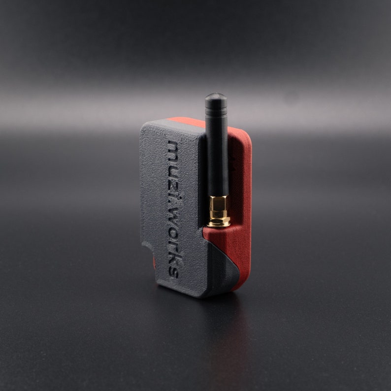 Pode incluir: Um pequeno dispositivo preto e vermelho com uma antena preta. O dispositivo tem o texto "muzi.works" impresso no lado.