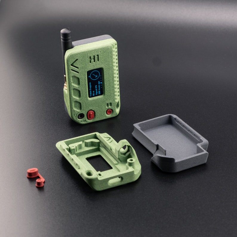 Pode incluir: Um dispositivo verde impresso em 3D com uma pequena tela e bot&otilde;es. O dispositivo est&aacute; em tr&ecirc;s partes, com um corpo principal, uma pe&ccedil;a superior separada e uma pe&ccedil;a lateral menor. O dispositivo tem uma pequena antena no topo.