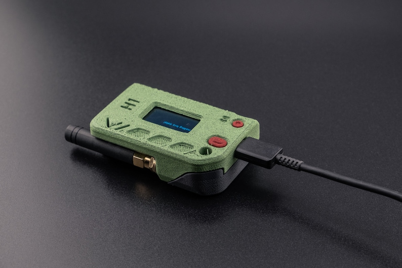 Pode incluir: Um dispositivo port&aacute;til verde e preto com uma pequena tela que exibe texto. O dispositivo tem um bot&atilde;o vermelho e uma antena preta. Um cabo preto est&aacute; conectado ao dispositivo.