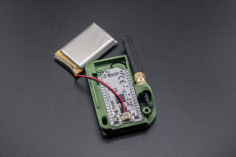 Pode incluir: Uma caixa verde impressa em 3D com uma pequena placa de circuito dentro. A caixa tem uma antena preta e uma bateria conectada &agrave; placa. O texto "HELtec" &eacute; vis&iacute;vel na placa.