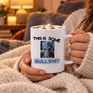 May include: A white ceramic mug filled with a hot beverage topped with marshmallows. The mug features a graphic of a grey alien face centered between the black text THIS IS SOME and the blue text BULLSHIT printed on the front surface.