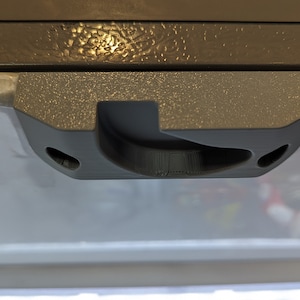 May include: A close-up of a gray plastic latch with a curved handle and two small holes on either side. The latch is attached to a white surface.