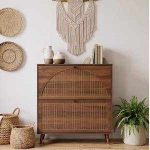 Puede incluir: Un mueble de madera oscura con diseño de listones y dos cajones. Encima, un tapiz de macramé y jarrones de cerámica. Cestas tejidas y un helecho en maceta completan la decoración bohemia.