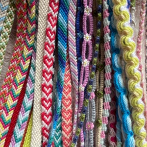 May include: A collection of colourful friendship bracelets made with different patterns and colours, including pink, blue, green, yellow, and white.