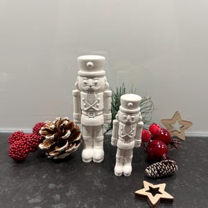 Könnte beinhalten: Zwei weiße Nussknacker-Figuren aus Keramik, eine große und eine kleine, stehen auf einer dunklen Oberfläche. Dekorative Elemente sind rote Beeren, ein Tannenzapfen und Holzsterne, die ein festliches Thema suggerieren. Die Figuren tragen traditionelle Uniformen.