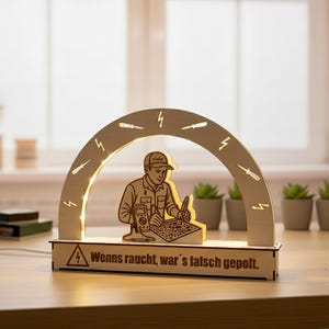 May include: A wooden light-up sign featuring an electrician figure inside an arch with tools and lightning bolts. The sign has the text "Wenns raucht, war's falsch gepolt." The sign is on a wooden table.
