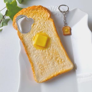 May include: A novelty bottle opener in the shape of a slice of toast with a pat of butter, and a matching keychain. The opener is silver and has a heart-shaped cutout. The keychain has a small toast charm with a butter square.