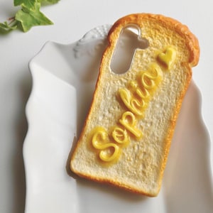 Może przedstawiać: Etui na telefon w kształcie kromki tostowego chleba, z napisem "Sophia" w żółtych, maślanych literach. Etui ma wycięcie na aparat i małe serce. Tost leży na białym talerzu.
