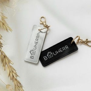 Könnte beinhalten: Zwei rechteckige Schlüsselanhänger, einer weiß und einer schwarz, mit goldfarbenen Verschlüssen. Jeder Schlüsselanhänger trägt den Text "BOUHERRIN" und ein Haus-Symbol mit einem Herzen. Die Schlüsselanhänger werden auf einer weißen Oberfläche präsentiert.