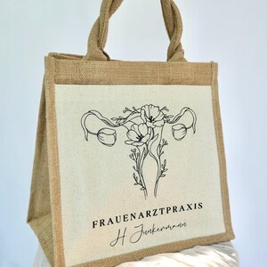 May include: A brown jute tote bag with a white canvas insert. The canvas insert features a black line drawing of a uterus with flowers. The text "FRAUENARZTPRAXIS H. Junkermann" is printed below the drawing.
