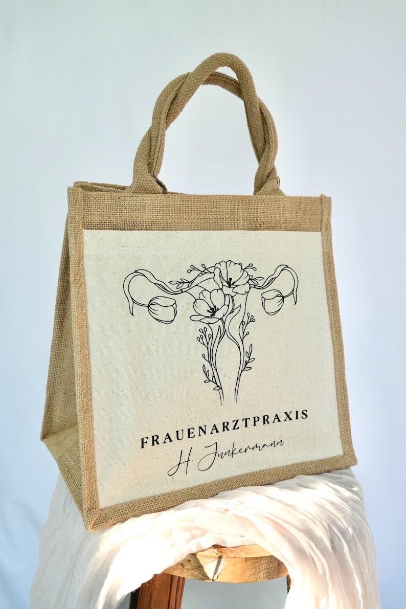 May include: A brown jute tote bag with a white canvas insert. The canvas insert features a black line drawing of a uterus with flowers. The text "FRAUENARZTPRAXIS H. Junkermann" is printed below the drawing.