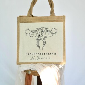 May include: A brown jute tote bag with a white canvas panel featuring a black line drawing of a uterus with flowers. The text "FRAUENARZTPRAXIS H Junkermann" is printed below the image.