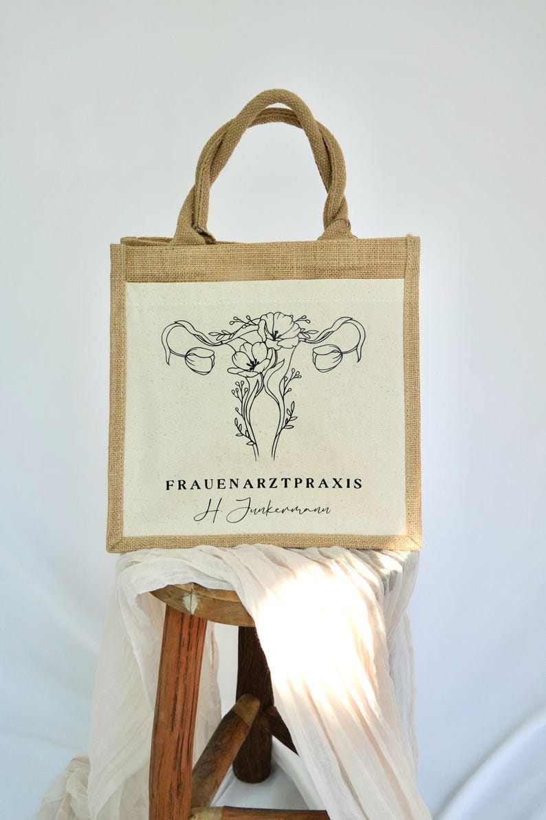 May include: A brown jute tote bag with a white canvas panel featuring a black line drawing of a uterus with flowers. The text "FRAUENARZTPRAXIS H Junkermann" is printed below the image.