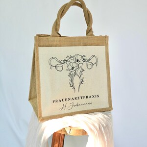 May include: A brown jute tote bag with a white canvas panel featuring a black line drawing of a uterus with flowers. The text "FRAUENARZTPRAXIS H Junkermann" is printed below the drawing.
