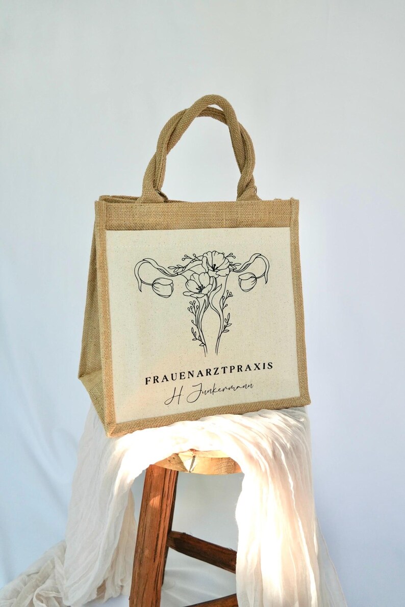 May include: A brown jute tote bag with a white canvas panel featuring a black line drawing of a uterus with flowers. The text "FRAUENARZTPRAXIS H Junkermann" is printed below the drawing.