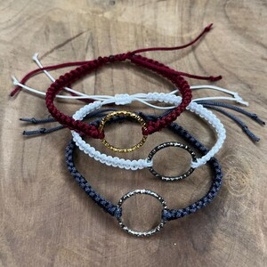 Puede incluir: Tres pulseras trenzadas con cordones de diferentes colores y dijes circulares de metal. Las pulseras son de color burdeos, blanco y gris oscuro.