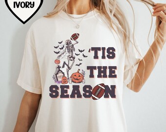May include: Ivory t-shirt with a graphic of a skeleton playing football with pumpkins and bats. The text 'Tis the Season' is printed on the shirt.