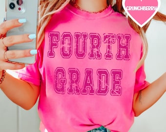 Puede incluir: Una camiseta rosa con el texto "FOURTH GRADE" en una fuente de estilo vintage y desgastada. El texto está en un color rosa más oscuro.