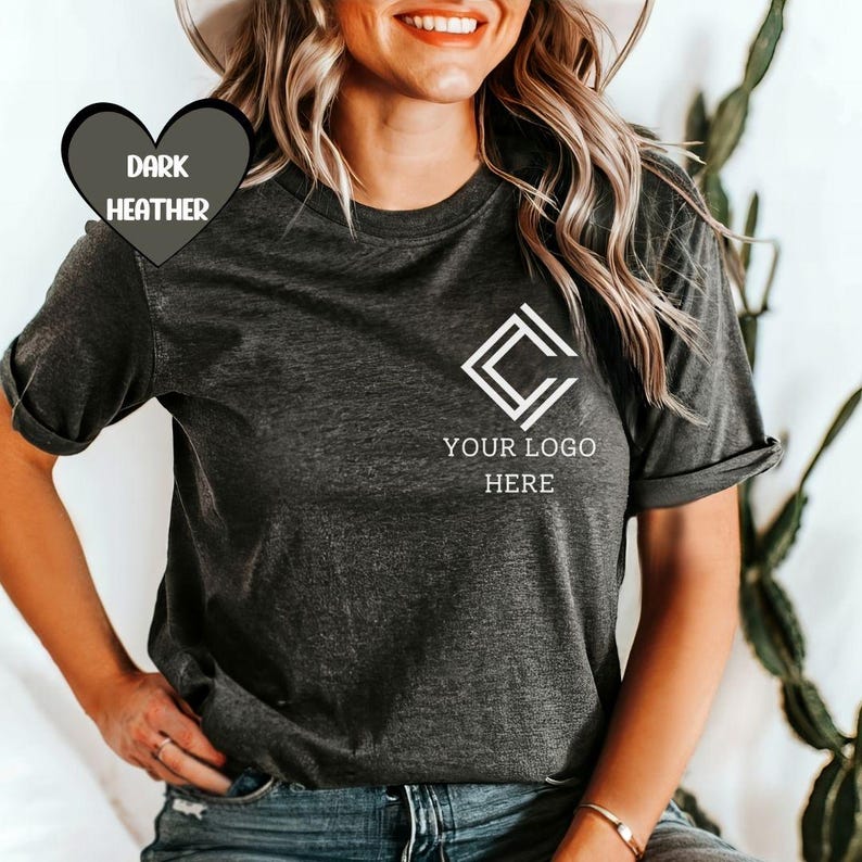 K&ouml;nnte beinhalten: Dunkelgraues meliertes T-Shirt mit wei&szlig;em Logo und dem Text "YOUR LOGO HERE". Das T-Shirt hat hochgekrempelte &Auml;rmel. Die Worte "DARK HEATHER" befinden sich in einer herzf&ouml;rmigen Grafik. Getragen von einer Person.