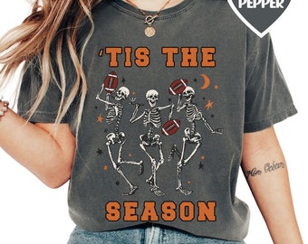 May include: A gray t-shirt with a vintage-style graphic of skeletons playing football. The text "'Tis the Season" is printed in orange on the shirt.