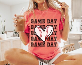 May include: A pink t-shirt with a distressed black graphic that says "GAME DAY" repeated multiple times. A red baseball with white stitching is in the center of the graphic, shaped like a heart with the number "12" inside.