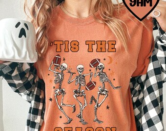 May include: Orange t-shirt with the text "'Tis the season" and a graphic of three skeletons playing football.