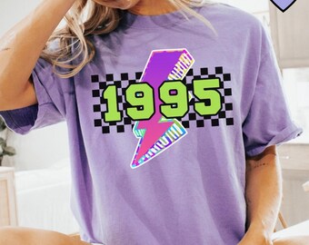 May include: A lilac-colored t-shirt featuring a graphic design with the year "1995" in neon green, set against a black and white checkered background. A pink and purple lightning bolt is centered below the numbers.