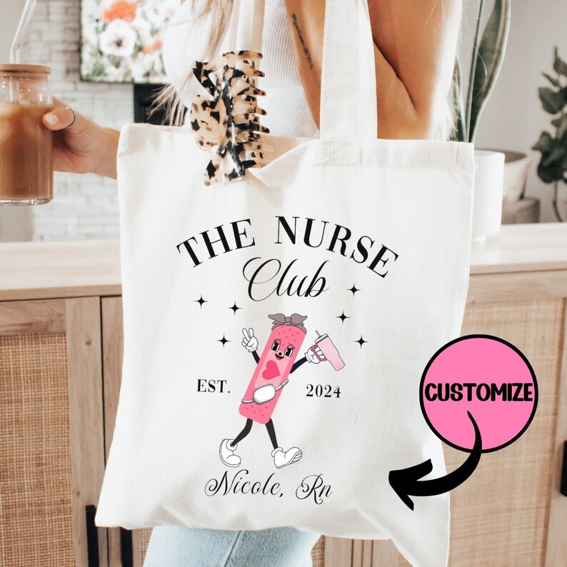 May include: White canvas tote bag with black text that reads "The Nurse Club" with a cartoon band-aid character holding a drink. The text "Est. 2024" is below the character. The text "Nicole, RN" is below the character. A pink circle with the word "Customize" is in the upper right corner of the image.