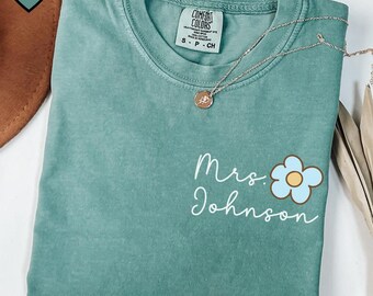 May include: Seafoam green Comfort Colors t-shirt with the text "Mrs. Johnson" and a light blue flower graphic. A silver necklace with a small pendant rests on the shirt. The shirt is size S.