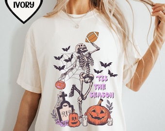 May include: Ivory t-shirt with a skeleton holding a football and a pumpkin. The skeleton is standing on a spiderweb and there is a tombstone that says "RIP" in the background. The text "Tis the Season" is written in purple on the shirt.