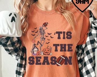 May include: Orange t-shirt with a skeleton holding a football and a pumpkin. The text on the shirt says "'Tis the Season" with a football graphic.