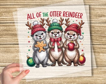 Könnte beinhalten: DTF-Transfer mit drei Cartoon-Ottern, die als Rentiere verkleidet sind, Hüte tragen und Weihnachtsschmuck halten. Der Text "ALL OF THE OTTER REINDEER" steht über den Ottern. Die Wörter "CUSTOM DTF TRANSFER" und "READY TO PRESS" sind ebenfalls sichtbar.