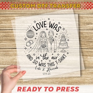 May include: A clear, ready-to-press DTF transfer with a wedding theme. The design features a bride and groom, a cake, rings, and the text "LOVE WAS IN THE AIR AND SO WAS THIS T-SHIRT" with the names Luke & Hannah and the date 12.10.25.
