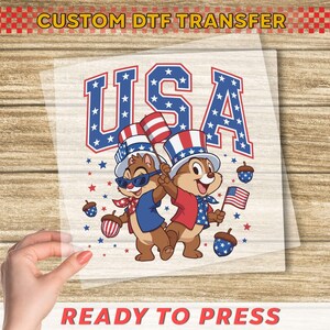 May include: A DTF transfer featuring two cartoon chipmunks in patriotic outfits, holding flags and wearing hats with the word "USA" in a star-spangled design. The text "CUSTOM DTF TRANSFER" and "READY TO PRESS" are also visible.