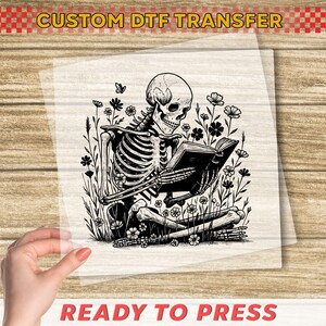 May include: A black and white DTF transfer featuring a skeleton reading a book while surrounded by flowers. The text "CUSTOM DTF TRANSFER" is at the top, and "READY TO PRESS" is at the bottom.