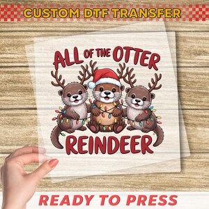 May include: A DTF transfer featuring three cartoon otters with reindeer antlers and Christmas lights. The otters are wearing Santa hats and the text reads "ALL OF THE OTTER REINDEER". The top text says "CUSTOM DTF TRANSFER" and the bottom text says "READY TO PRESS".