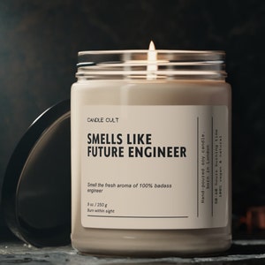 Pode incluir: Uma vela de soja bege com um rótulo preto que diz "CANDLE CULT", "SMELLS LIKE FUTURE ENGINEER", "Smell the fresh aroma of 100% badass engineer", "9 oz./250 g", "Burn within sight", "Hand-poured soy candle. born in London", "50-60 hours burning time", "100% vegan & natural". A vela está acesa e a chama é visível.