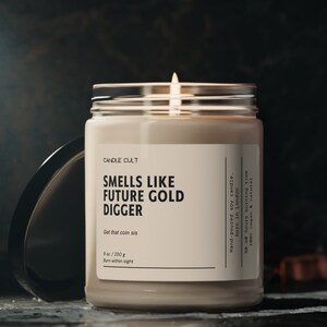 May include: A beige candle in a clear glass jar with a black lid. The label reads "CANDLE CULT", "SMELLS LIKE FUTURE GOLD DIGGER", "Get that coin sis", "9 oz./250 g", and "Burn within sight". The candle is hand-poured soy and has a burn time of 50-60 hours. It is 100% vegan and natural.
