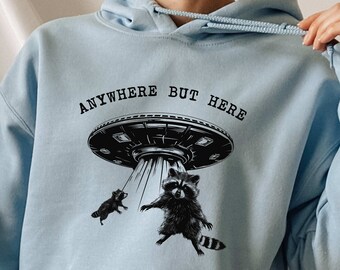 Peut inclure: Sweat-shirt à capuche bleu clair avec le texte "Anywhere But Here" et une illustration en noir et blanc d'un OVNI avec deux ratons laveurs qui en pendent.
