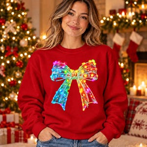 Puede incluir: Una sudadera roja de manga larga con un gráfico central grande de un lazo translúcido decorado con luces navideñas brillantes y multicolores en tonos azul, verde, amarillo y rosa sobre la tela.